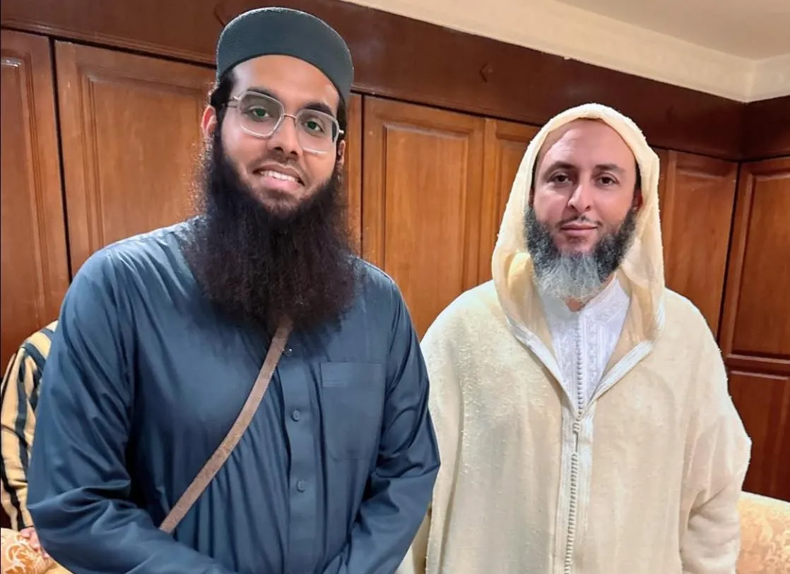 الشيخ سعيد الكملي والشيخ شمائل الندوي معاً صورة للشيخ سعيد الكملي (يمين) والشيخ شمائل الندوي (يسار) يقفان جنباً إلى جنب في غرفة ذات خلفية خشبية، كلاهما يرتدي الزي الإسلامي التقليدي ويبتسمان بهدوء.